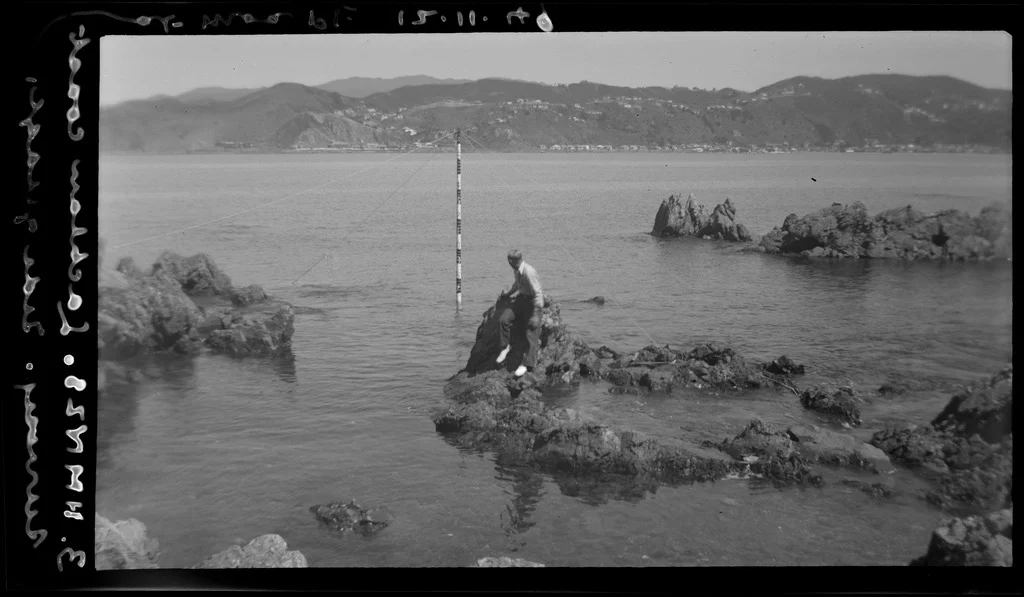 An archival photograph of a surveyor on coastal rocks beside a marked pole in shallow water