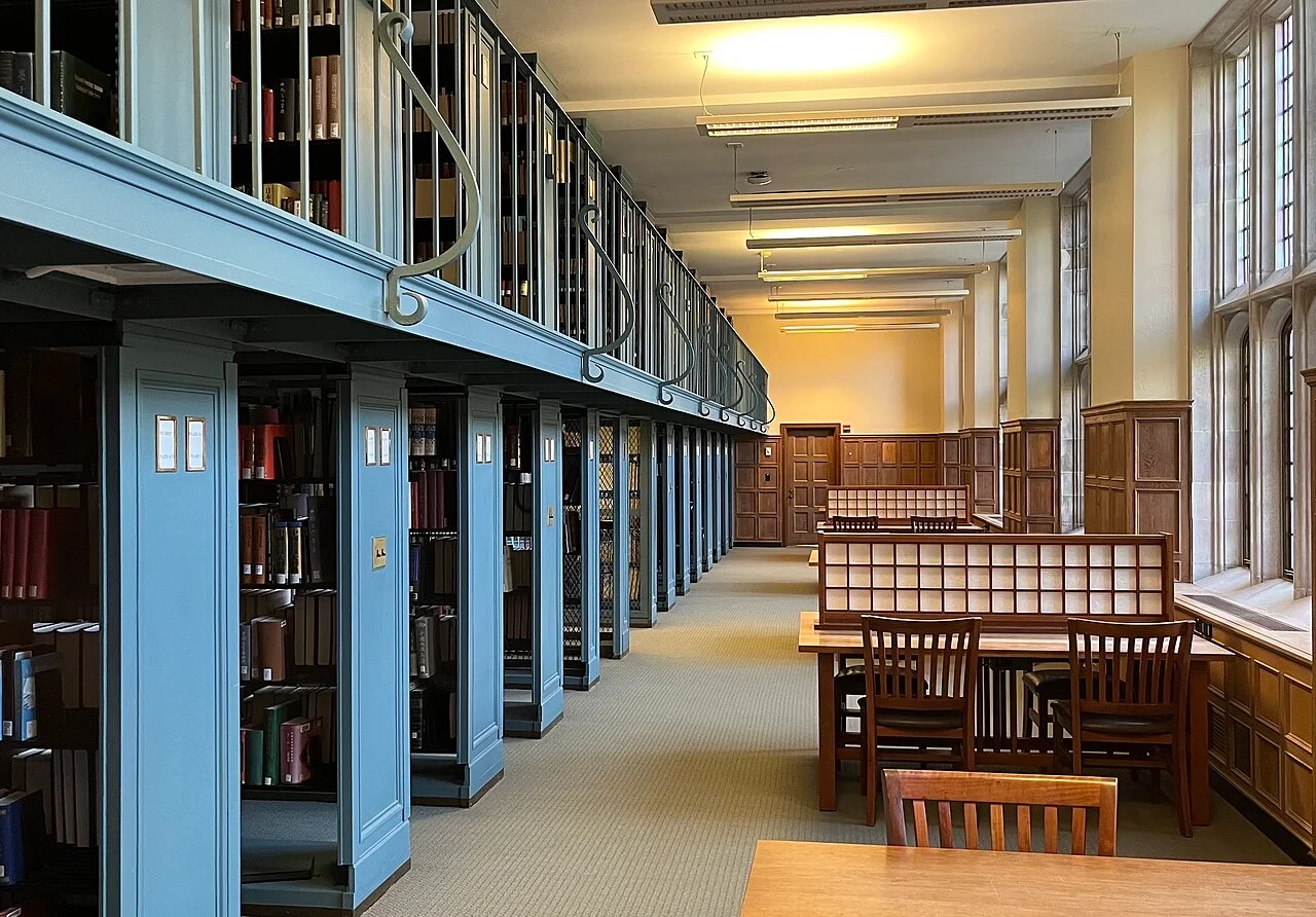 A library reading room with wooden tables between tall book stacks